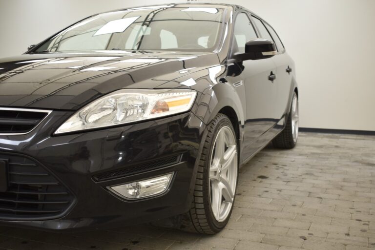 A black car parked on a tiled floor inside a well-lit, plain room. The front and side of the car are visible, showcasing its headlights and tires.