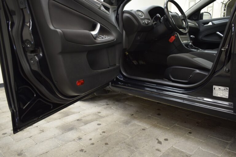 Open driver-side door of a black car showing the interior, including the driver's seat, steering wheel, and side door panel with control buttons. The car is parked on a tiled surface.