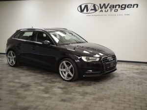 A black Audi hatchback is parked in a showroom with a tiled floor. The wall in the background has a Wangen Auto logo and the website address.